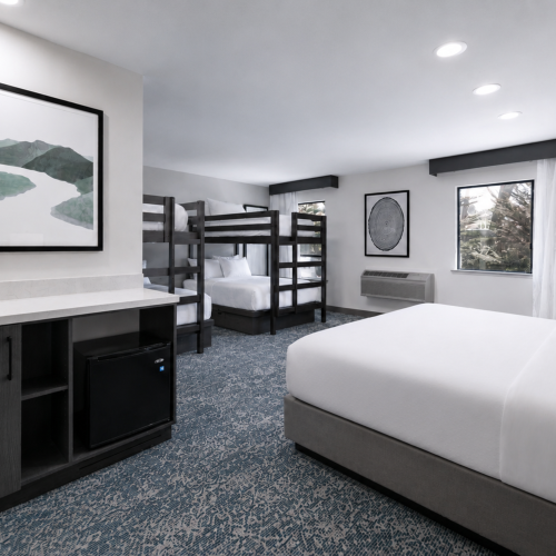 Modern hotel room with a sofa, bunk beds, windows, patterned carpet, and a vanity sink.