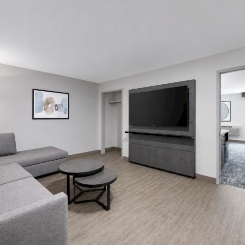 A modern living room with a gray sofa, round coffee table, and a wall-mounted TV beside an open doorway to a bedroom.