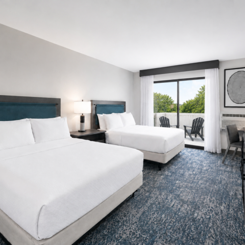 A bright hotel room with two white beds, blue headboards, a desk, window curtains, and an outdoor view.