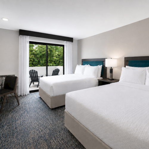 Bright hotel room with two beds, teal headboards, bedside lamps, and a window with chairs on a balcony, tidy and modern.