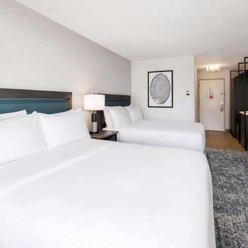 A modern hotel room with two white beds, bedside lamps, a dresser, wall art, a TV, and a bathroom doorway in the back.