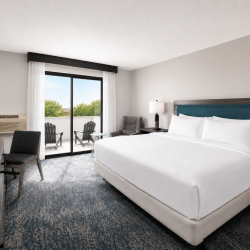 A modern hotel room with a large bed, bedside lamps, chairs by a glass door, and sheer curtains letting in daylight.