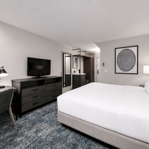A modern hotel bedroom with a white bed, TV on a dark console, framed wall art, a desk lamp, and a mirror.