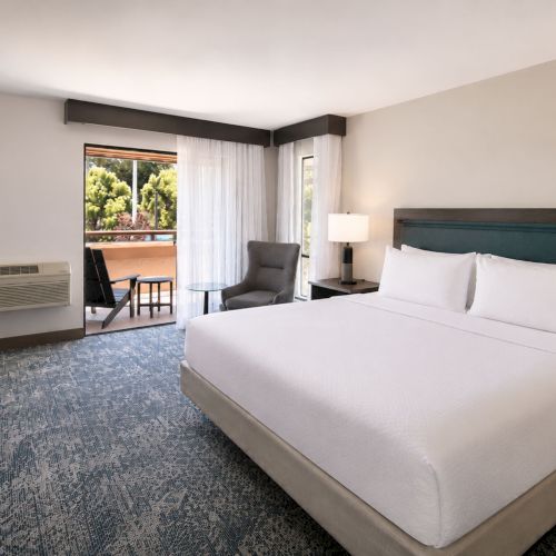 Modern hotel room with a king bed, bedside lamps, blue patterned carpet, and sheer curtains by a balcony door.