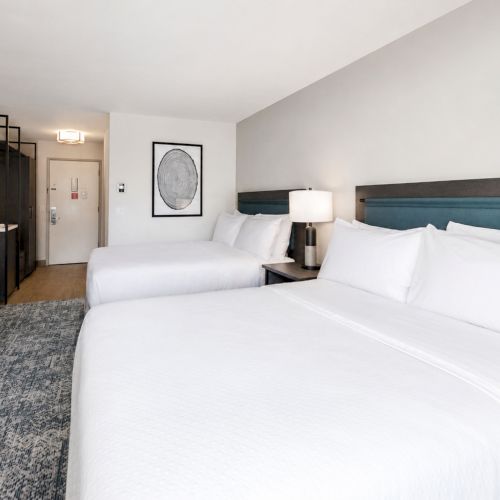 A modern hotel room featuring two white beds, a dark headboard, bedside lamps, framed art, and a dresser with a TV.