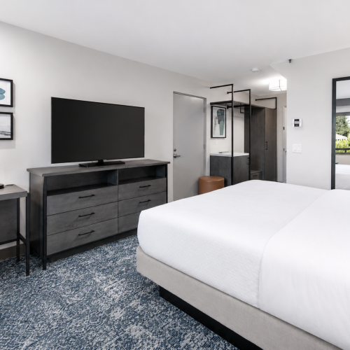 A modern hotel room with a white bed, wall-mounted TV, gray dresser, and framed art and mirrors in a bright setting.