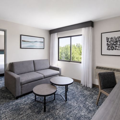 A modern hotel lounge with a gray sofa, round coffee tables, big window with sheer curtains, framed art, and an air conditioner.