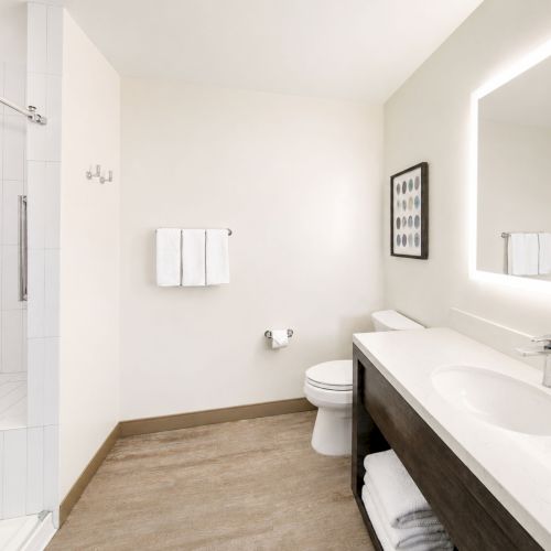 Bright modern bathroom with glass shower, tiled walls, glowing mirror, double vanity, toilet, towels, and metal fixtures.