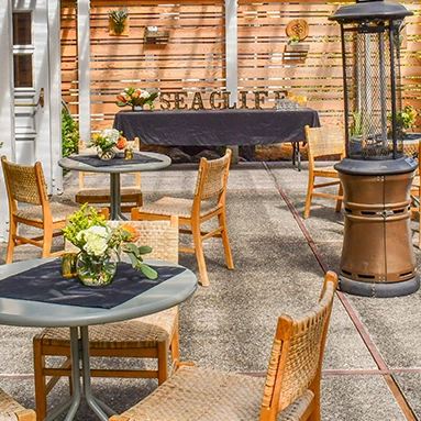 An outdoor patio with wooden chairs and tables, potted plants, a central fire pit, and an &ldquo;EAGLE&rdquo; sign in the background.