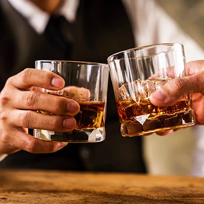 Two people clink glasses filled with amber whiskey, toasting together during a celebration.