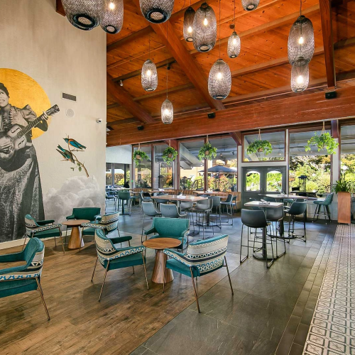 A modern caf&eacute; interior with teal chairs, hanging lantern lights, and a wall mural of a guitarist, plus indoor plants by the windows, serving.