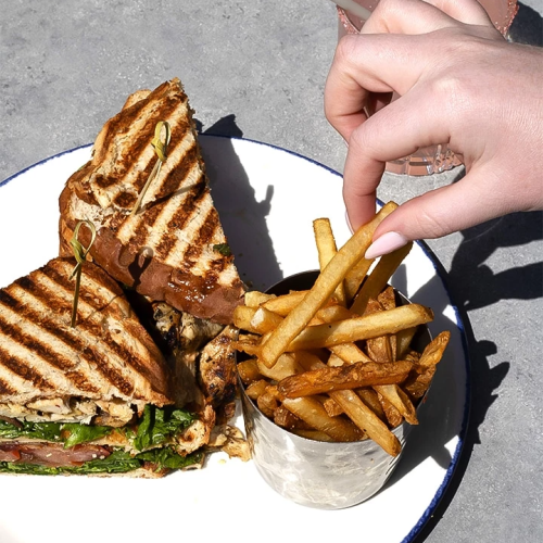 A hand adds fries to a plate beside a grilled sandwich and greens.