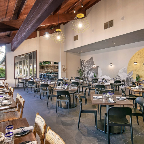 A bright restaurant dining room with neatly set tables, pendant lights, and a large bird mural on the wall.