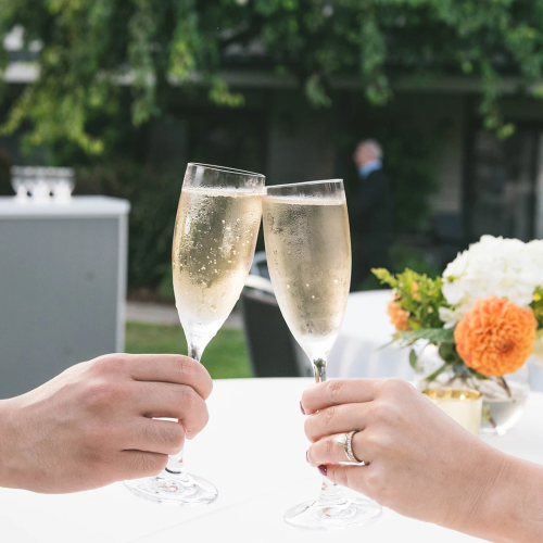 Two people clink champagne flutes on a sunny outdoor patio, with flowers on the table.
