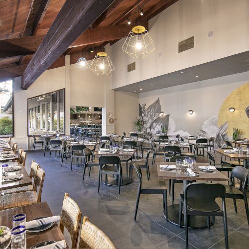 A modern restaurant dining room with set tables, hanging lights, and a large bird mural on the wall.
