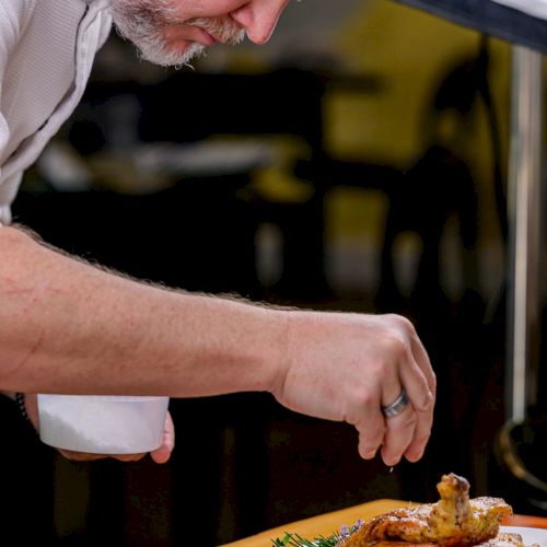 An older man leans over a wooden counter, seasoning or carving a roasted chicken while holding a white cup and smiling over his work.