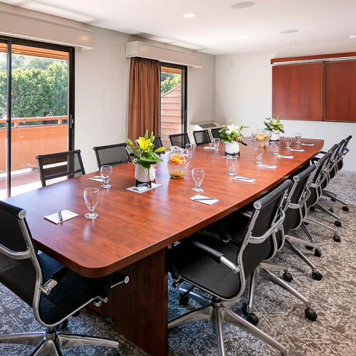 A modern conference room with a long wooden table, black rolling chairs, place settings, fresh flowers, windows with orange curtains, and a sideboard in the back.