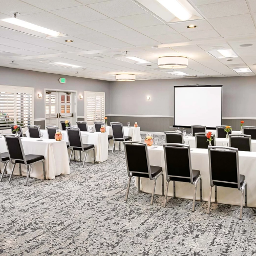 A conference setup with round tables and chairs arranged for a meeting, projector screen at the front, and floral centerpieces on each table.