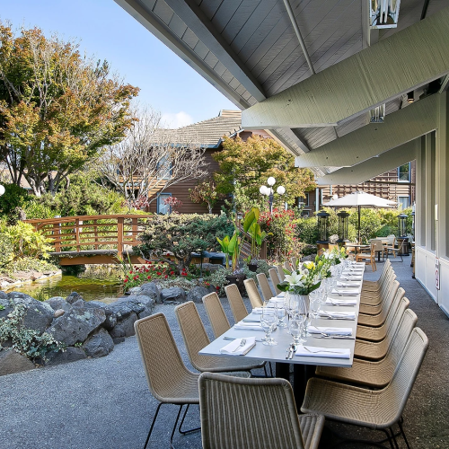 Outdoor dining setup along a covered patio with a long table, beige chairs, white napkins, and a garden with rocks, trees, and a wooden bridge in the background.