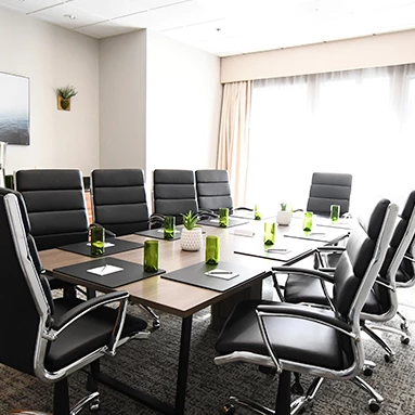 A modern conference room with a long table, black leather chairs, green bottled drinks, plant decor, a wall-mounted TV, cabinets, and large windows with beige curtains.