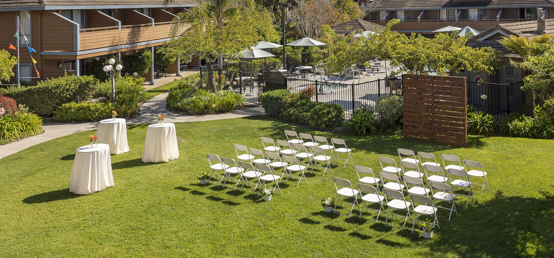 An outdoor garden wedding setup with rows of chairs, round tables, umbrellas, lush landscaping, and surrounding buildings under a clear sky.