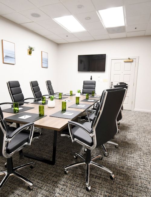 A bright office conference room with a long table, black chairs, framed photos, and a cabinet and TV on the wall.