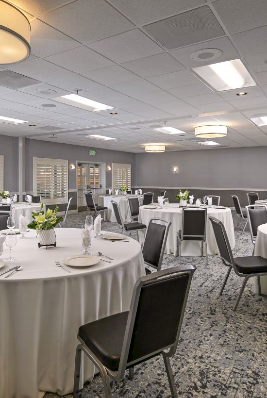 A banquet hall with round tables set for dinner, gray chairs, and bright ceiling lights, with an exit sign near the door.
