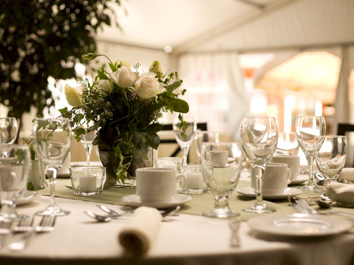 A banquet table is set with glassware and cups, with a bouquet of white roses as the centerpiece.