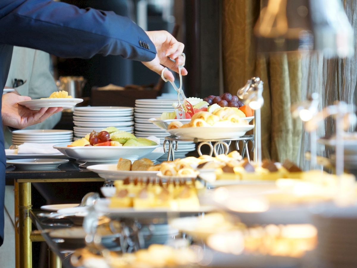 A server serves a fresh fruit salad at a catered buffet, holding tongs above stacked plates of food.