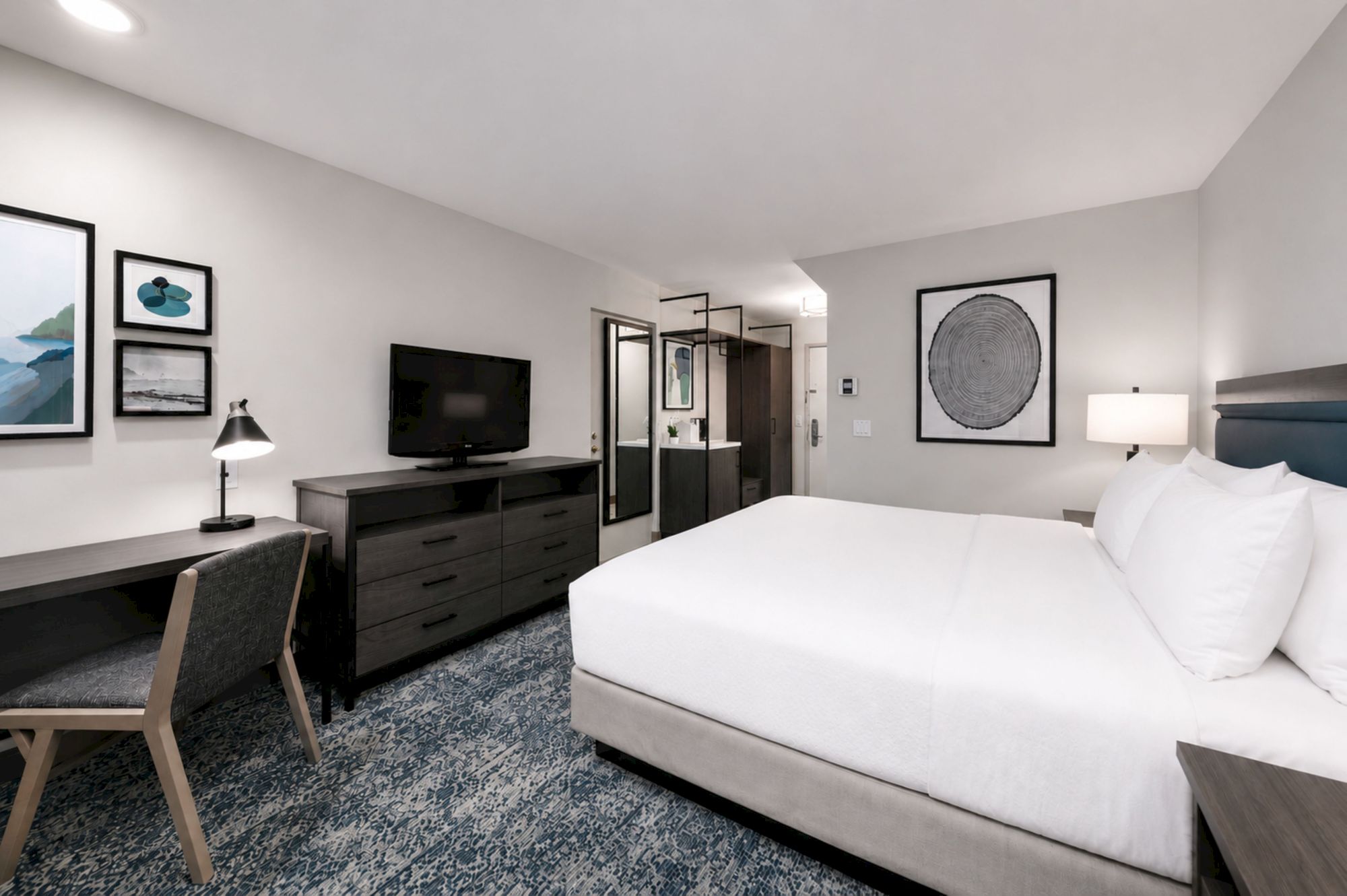 A modern hotel room: a white bed, TV on a dresser, framed wall art, a lamp, and dark glass-front shelving.