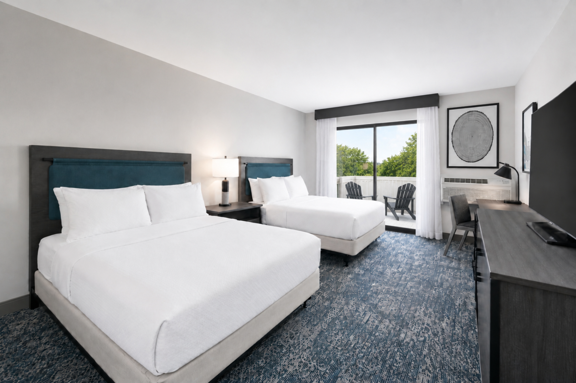 A bright hotel room with two large white beds, blue headboards, a window with curtains, a desk, and chairs overlooking trees.