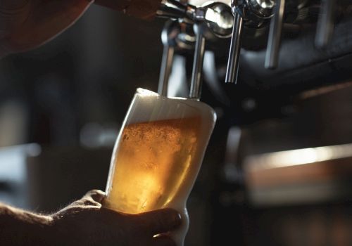 A bartender pours amber beer into a foamy glass mug with an out-of-focus bar behind it.