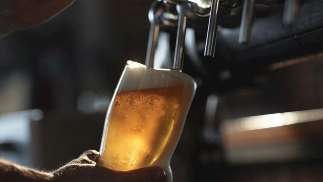 A bartender pours amber beer into a foamy glass mug with an out-of-focus bar behind it.