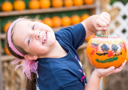 A smiling child holds a carved, decorated Halloween pumpkin at an outdoor pumpkin display, ready for trick-or-treating!.