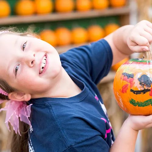 A smiling child holds a carved, decorated Halloween pumpkin at an outdoor pumpkin display, ready for trick-or-treating!.
