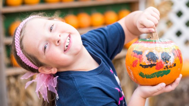 A smiling child holds a carved, decorated Halloween pumpkin at an outdoor pumpkin display, ready for trick-or-treating!.
